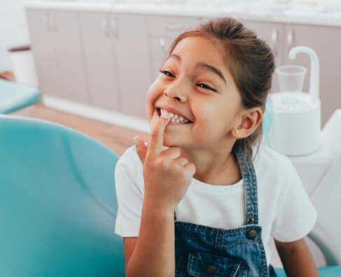 Young girl smiling in dental office, pointing to her tooth during checkup.