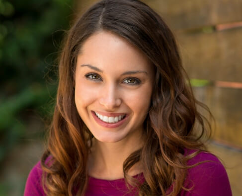 Woman smiling at camera wearing purple shirt, brick wall background for Breezy Point Smiles dental practice.