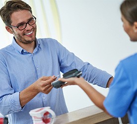 Man in blue shirt smiling while handing credit card to dental receptionist at office counter.