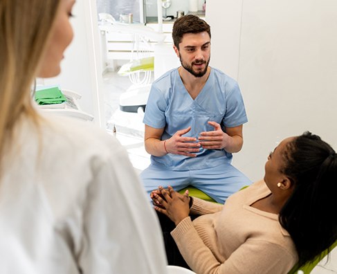 Healthcare provider in blue scrubs discussing treatment with two patients in dental office setting.