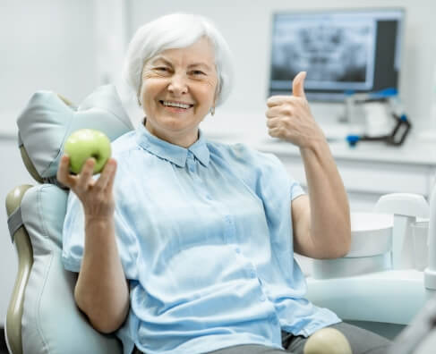Senior woman holding green apple giving thumbs up in dental office setting