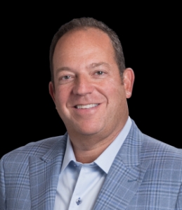 Professional headshot of a smiling man in light blue plaid blazer against black background.