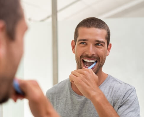 Man brushing teeth at bathroom mirror, smiling at reflection during morning routine.