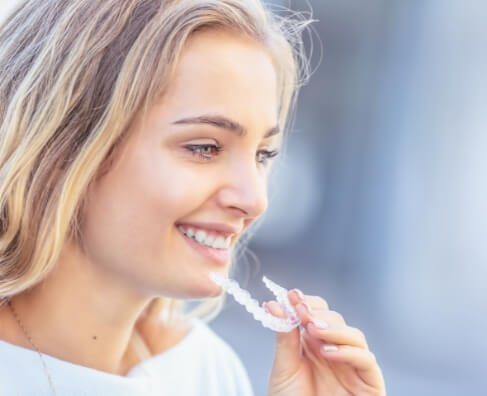Young woman smiling while holding clear dental aligners, bright outdoor setting.