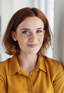 Woman in mustard yellow shirt smiling at camera in bright, professional office setting.