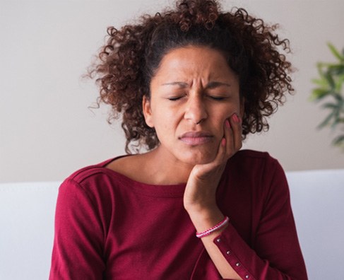 Woman in burgundy shirt looking concerned with hand on face, sitting indoors.