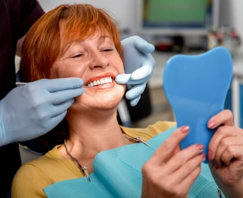 Woman receiving dental examination in dentist's office, holding mirror while dentist checks teeth.