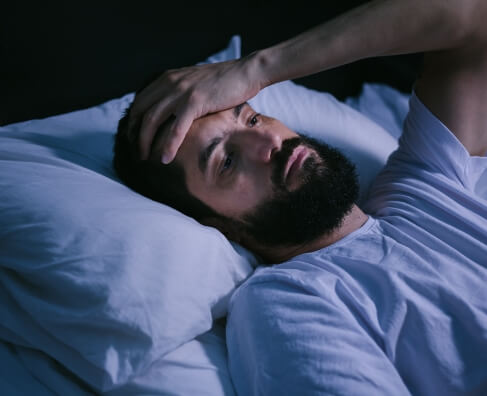 Man lying in bed at night, appearing distressed or unable to sleep, holding head.