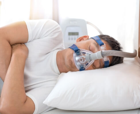 Man sleeping in bed wearing sleep apnea mask and white pajamas.