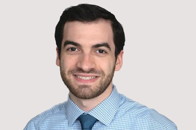 Professional headshot of smiling man in blue checkered shirt and tie against neutral background.